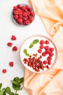 Yogurt with raspberry and goji berries in ceramic bowl on white concrete background and orange