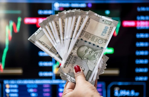 A person holding Indian 500 rupee currency bank note against share market graph on a display