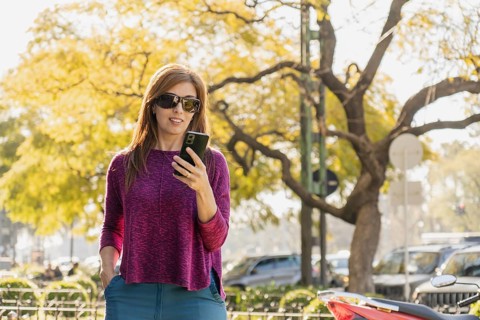 Stylish middle-aged woman smiles as she looks at her smartphone in a park at sunset. Wearing