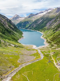 An alpine lake surrounded by green meadows and mountains, with a river and a hiking trail under a
