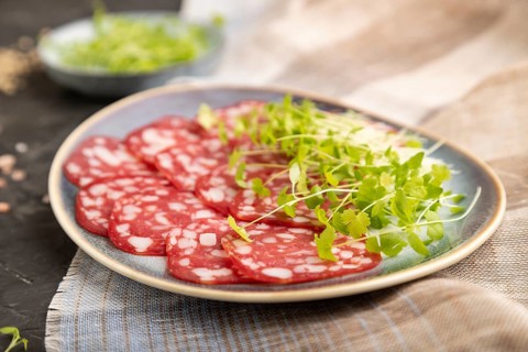 Slices of smoked cervelat salami sausage with spinach microgreen, salt and pepper on black concrete