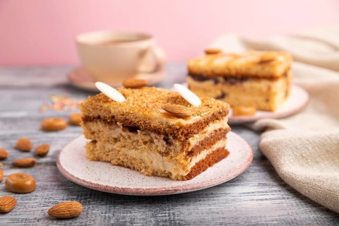 Honey cake with milk cream, caramel, almonds and a cup of coffee on a gray and pink background and