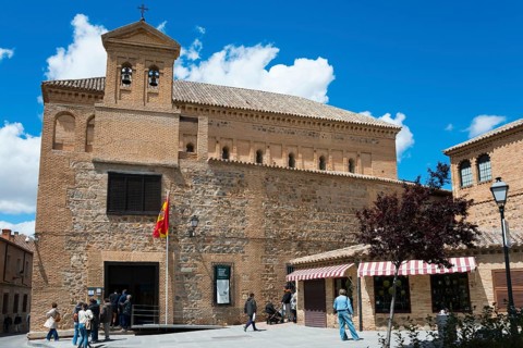 Historic church in Toledo with flag, people strolling and beautiful blue sky and trees in the
