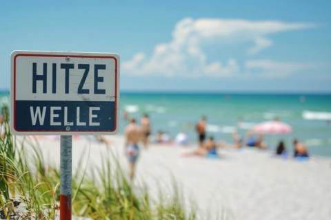 Warning sign with German text 'Hitzewelle' (Heat wave) in front of blurry sunny beach with people.
