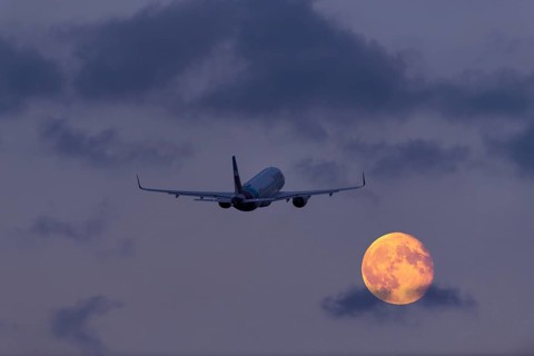 An aeroplane flies through the cloudy sky at night in front of a large, shining full moon, super