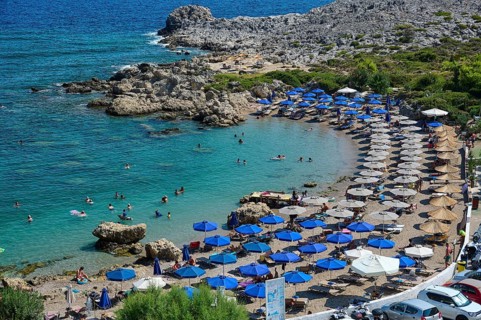 Beach full of deckchairs and blue umbrellas, lots of people in and around the clear water,