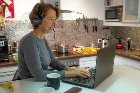 Woman, mid-50s, works from home, with laptop and communicates with colleagues via headset, home