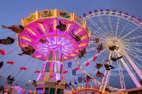 A lively funfair at dusk, illuminated by a large Ferris wheel and a colourful chain carousel,