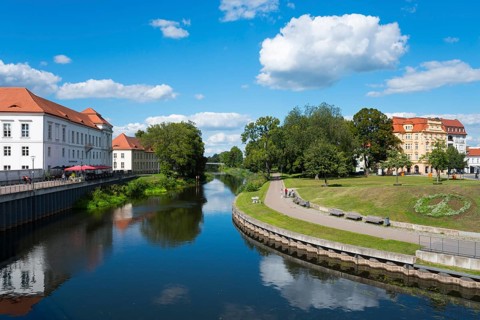 A quiet river with trees, park benches and historic buildings under a blue sky, Oranienburg, Havel