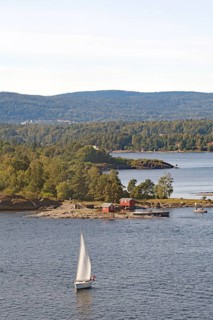 A single sailboat sails peacefully on the water, near a green island and wooded hills, Oslofjord,