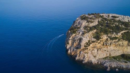 Drone shot, boat sailing along a rocky cliff over the clear blue sea, Anthony Quinn Bay, Vagies