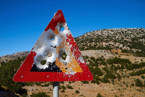 A damaged road sign with bullet holes in front of a mountainous landscape and blue sky, Lefka Ori,