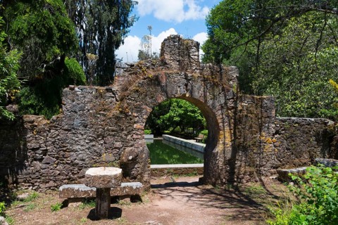 Ein alter Steinbogen führt in einen grünen Garten an einem sonnigen Tag, Mata Nacional do Buçaco,