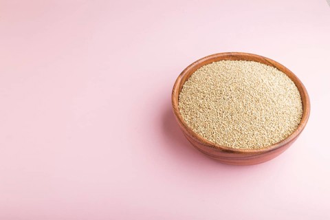 Wooden bowl with raw white quinoa seeds on a pastel pink background. Side view, copy space