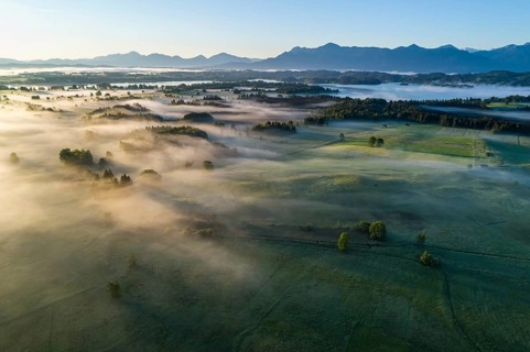Aerial view of meadows and trees in front of mountains in backlight, sunrise, fog, autumn, Murnau,