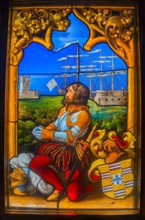 Stained glass window shows a knight in armour with a sailing ship and castles in the background,