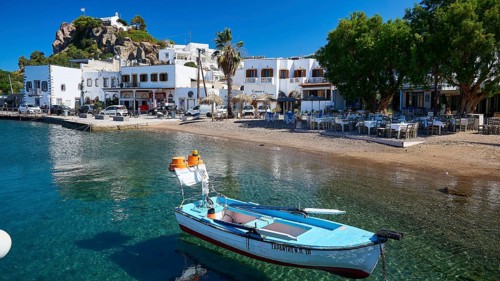 A fishing boat lies on the shore of a Greek island, next to cafés and historic buildings along the