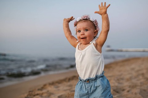 A baby in summery clothes and flowers in her hair stands on the beach at sunset and stretches both