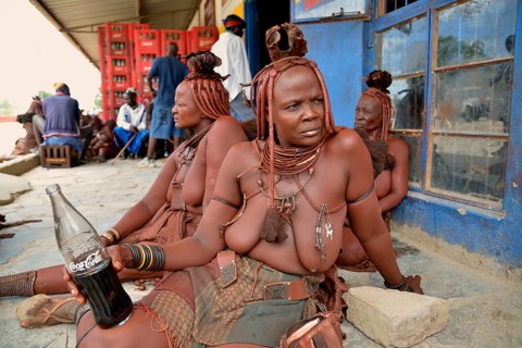Traditionally dressed Himba woman drinking a coke in front of a bar, Opuwo, Kaokoland, Kunene,