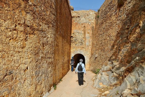 Two people walking through a narrow passageway between high stone walls of an ancient fortress,