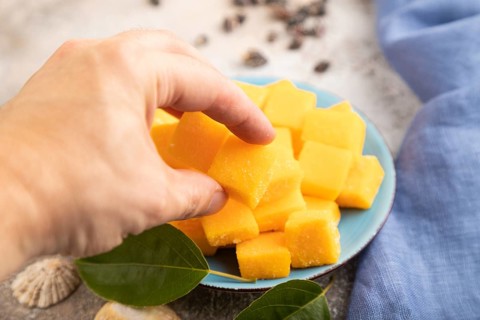 Dried and candied mango cubes on blue plate with hand on brown concrete background and linen