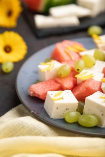 Vegetarian salad with watermelon, feta cheese, and grapes on blue ceramic plate on black concrete