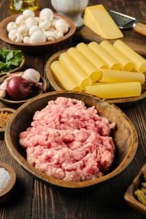 A wooden bowl filled with raw ground meat is surrounded by pasta, mushrooms, garlic, and cheese on