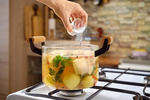 A hand adds vodka to a pot with fish soup, vegetables and herbs on a stove. The kitchen features a