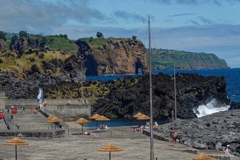 A rocky coastline with people in a seawater swimming pool resting under parasols while the sea