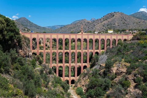 An old brick aqueduct spans a wooded ravine against a mountain backdrop under a clear blue sky,
