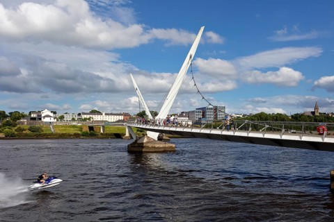 A jet ski passes under a striking bridge surrounded by calm waters, Londonderry