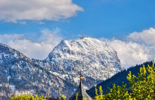 Snow-covered mountain peak under a blue sky with clouds, in the foreground a church tower and green
