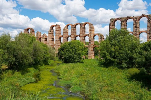 Antike Aquädukt-Ruinen inmitten von üppiger Vegetation und Fluss unter blauem Himmel, Acueducto de