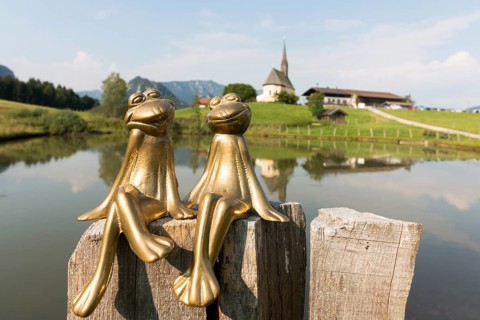 Two brass frogs, behind them the church of St Nicholas, Einsiedl, Inzell, Bavaria, Germany, Europe