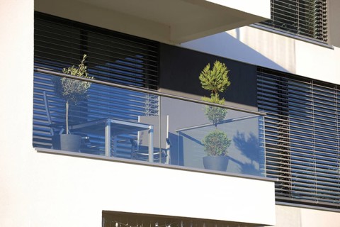 Balcony railing made of glass and stainless steel, behind it windows with modern blinds