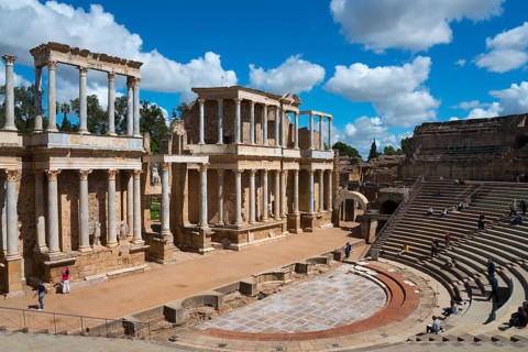 Wide view of a Roman theatre with ancient ruins and spectators on the steps under a blue sky,