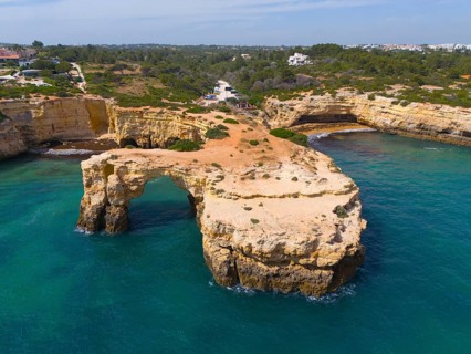 Large, rocky coastline with a natural arch surrounded by clear water and green vegetation, aerial