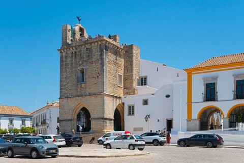 Historic church with bell tower, surrounded by cars and white buildings, in sunny weather, Sé
