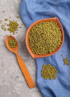 Clay bowl with raw green mung bean and wooden spoon on a gray concrete background and blue textile.