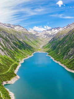 Landscape with a long, blue lake surrounded by high grass and tree-covered hills and snow-capped