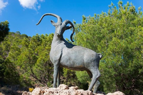Bronze statue of an ibex in nature on a rock base under a blue sky, Área Recreativa de Sedella,