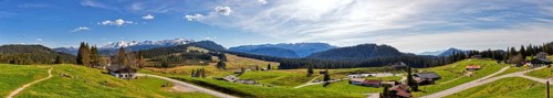 Wide green meadows in front of a mountain range, dotted with huts, under a partly cloudy blue sky,