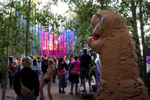 A festival visitor dressed as a dinosaur in front of a stage in the forest at the Melt Festival in