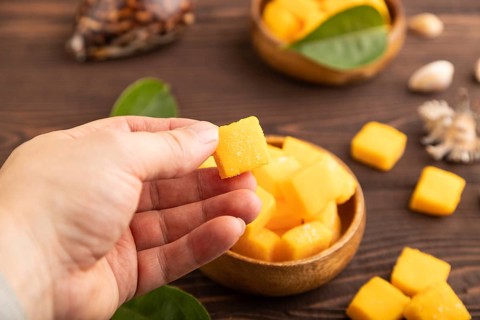 Dried and candied mango cubes in wooden bowls on brown wooden textured background with hand. Side