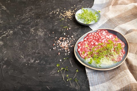 Slices of smoked cervelat salami sausage with spinach microgreen, salt and pepper on black concrete