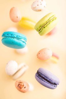 Multicolored flying macaroons and chocolate eggs frozen in the air on blurred beige background. top