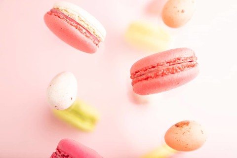 Multicolored flying macaroons and chocolate eggs frozen in the air on blurred pink background. top