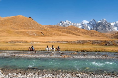 Riders riding through river in front of mountain landscape with yellow meadows, Kol Suu River and