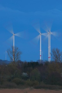 Wind energy systems turn in the evening light over a landscape framed by trees, wind turbines in