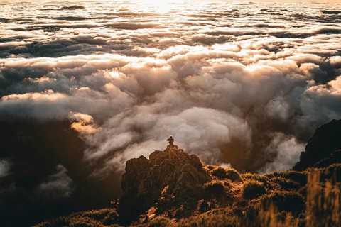 Person sitting on a rock looking over a cloud cover in the golden light of sunset, Pico Ruivo,